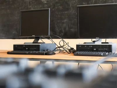 Zwei Computer stehen auf einem Tisch, im Hintergrund ist eine Tafel zu sehen.