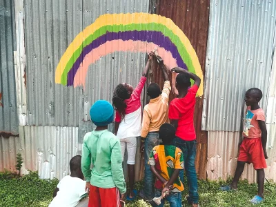 Einige Kinder malen einen bunten Regenbogen an den Zaun.