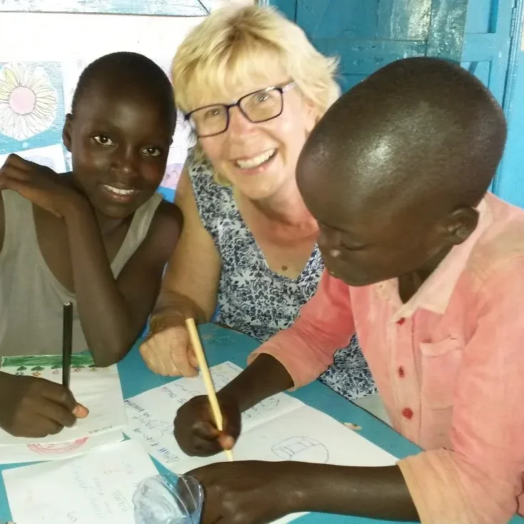 Regine Mazitschek mit zwei Malaika-Kindern beim gemeinsamen Zeichnen im Klassenzimmer, alle drei lachen in die Kamera