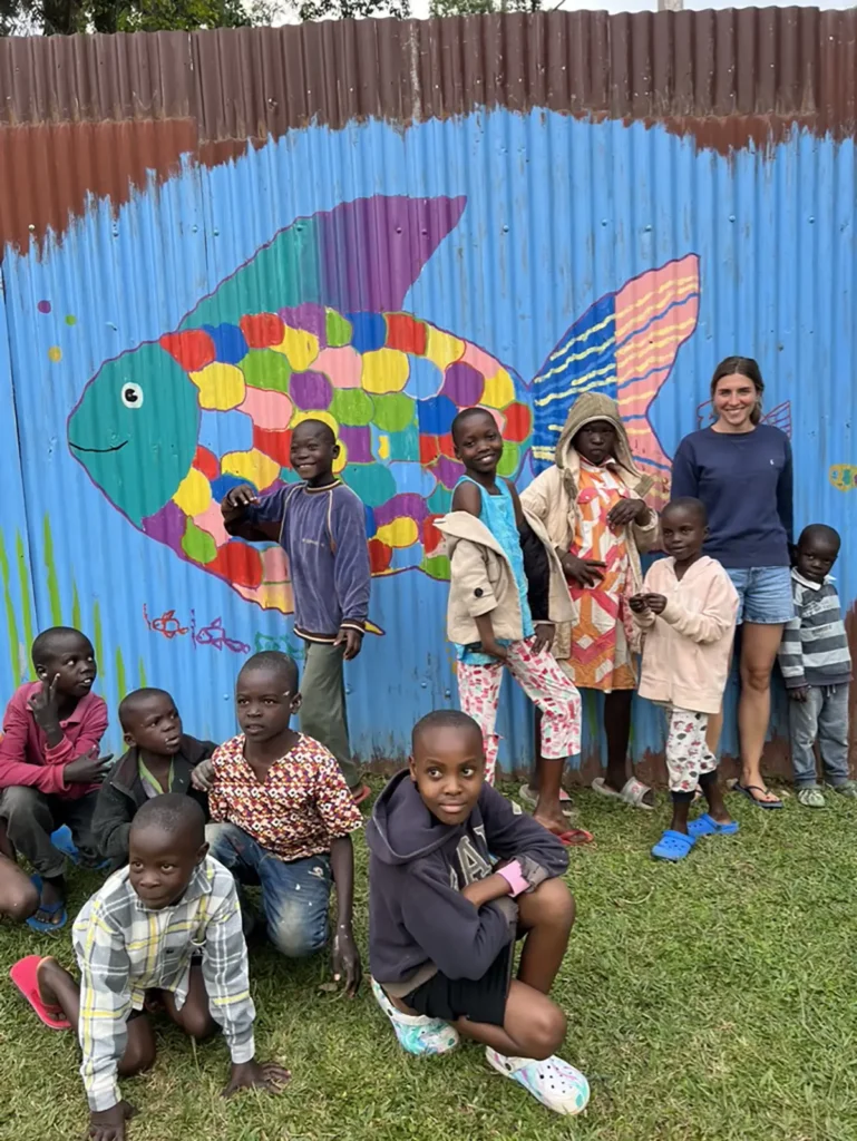Laura steht mit einer Gruppe von Kindern vor dem buten Regenbogenfisch der auf den Zaun gemalt ist