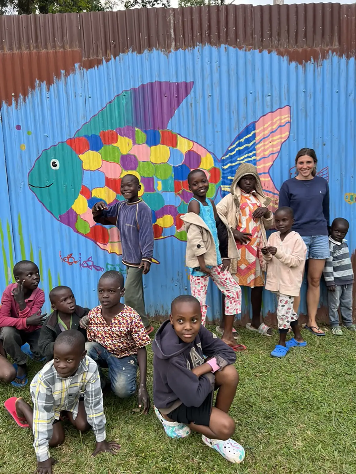 Laura steht mit einer Gruppe von Kindern vor dem buten Regenbogenfisch der auf den Zaun gemalt ist