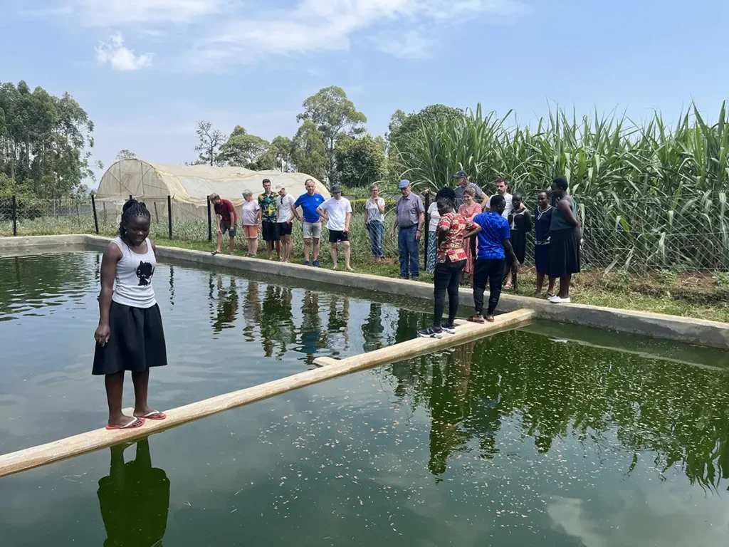 Malaika-Mitarbeitende und Besucher stehen gemeinsam am Fischteich und besichtigen die eigene Tilapia-Zucht des Malaika Children’s Home.