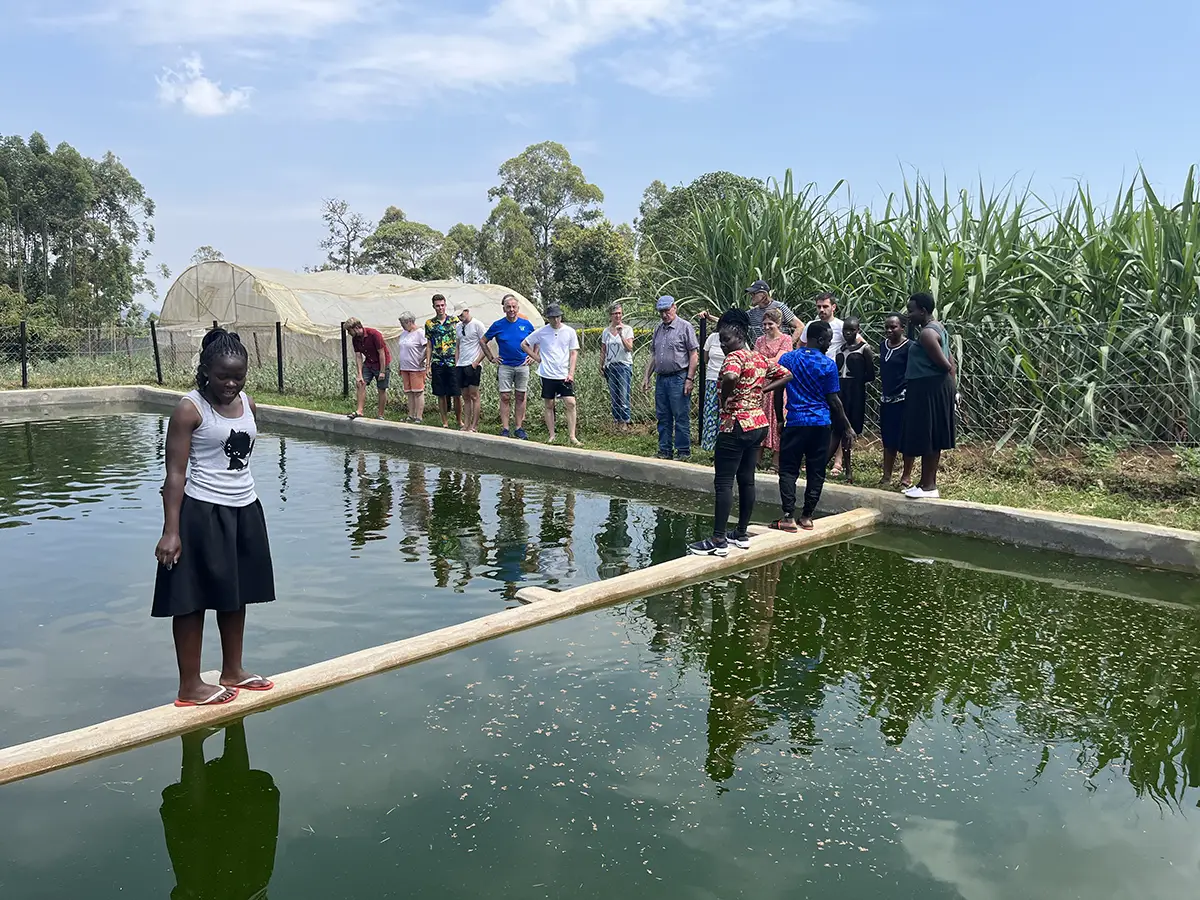 Malaika-Mitarbeitende und Besucher stehen gemeinsam am Fischteich und besichtigen die eigene Tilapia-Zucht des Malaika Children’s Home.