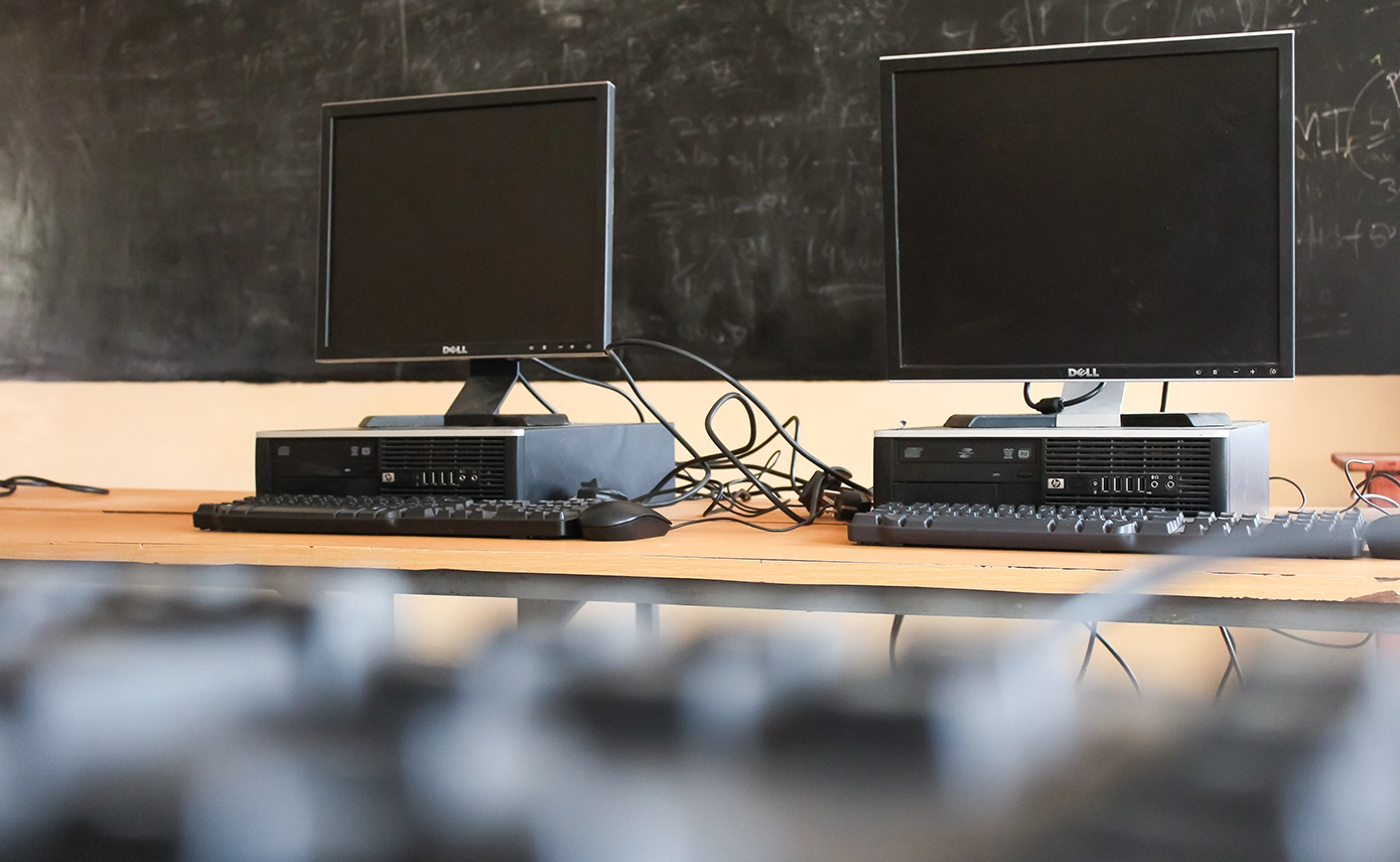 Zwei Computer stehen auf einem Tisch, im Hintergrund ist eine Tafel zu sehen.